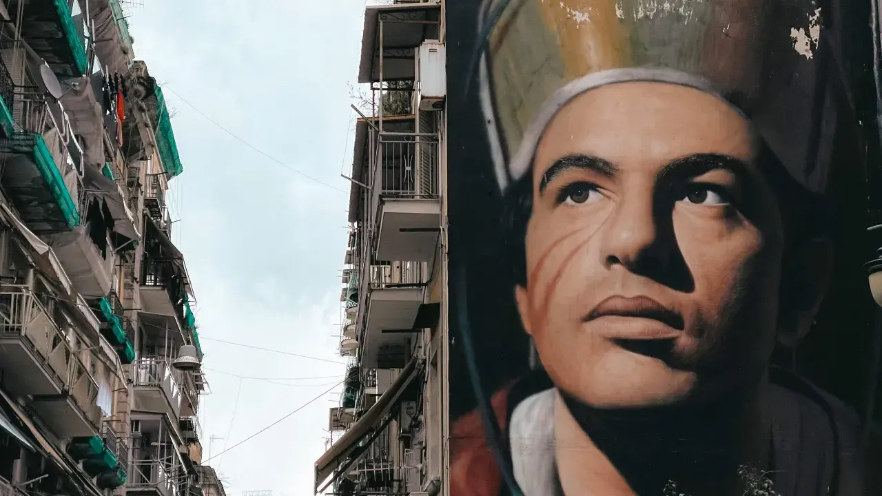Un imponente murales napoli raffigura un volto intenso, con un cappello particolare. Accanto, palazzi con balconi e panni stesi.