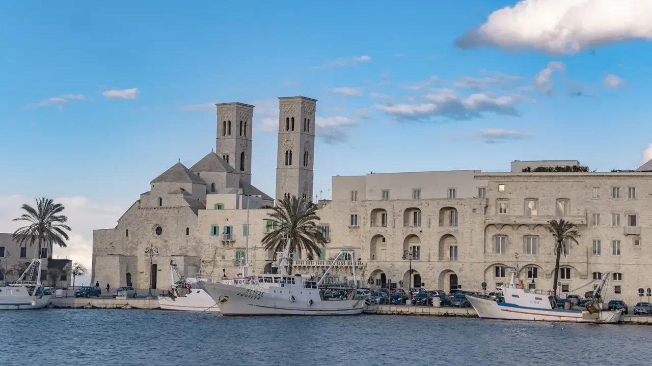Molfetta cosa vedere: la Basilica e il porto con pescherecci sotto un cielo azzurro e nuvoloso.