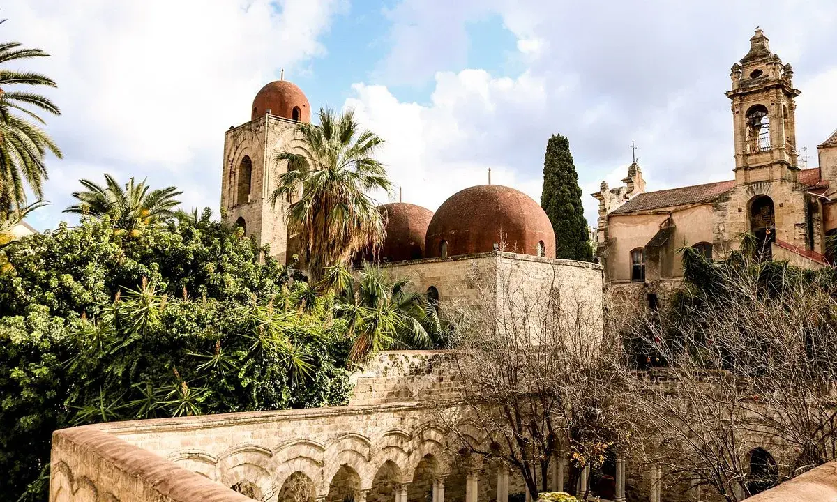 Le iconiche cupole rosse di Palermo si stagliano tra palme e vegetazione lussureggiante, un'antica bellezza sotto un cielo nuvoloso.