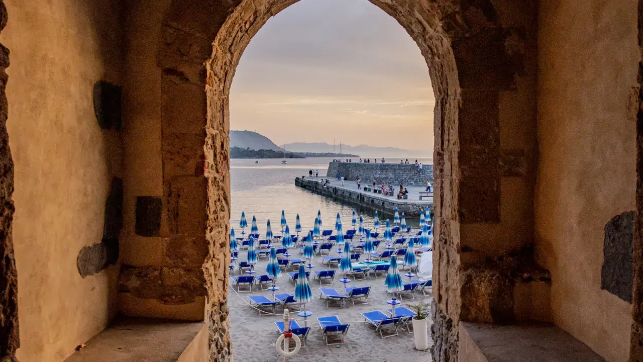 Arco antico incornicia la spiaggia di Cefalù con ombrelloni blu e lettini, vista sul mare e sul molo affollato.