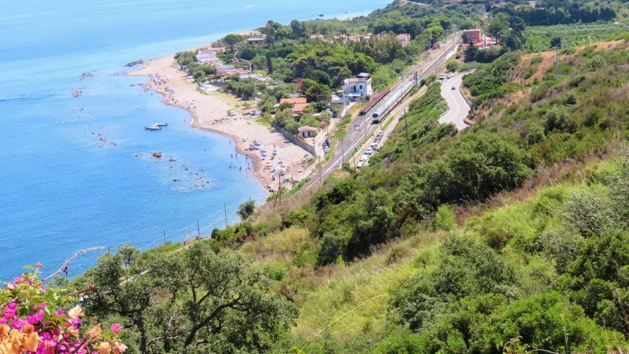 Spiaggia turchina finale di Pollina, con bagnanti e barche. Sopra, una ferrovia e una strada serpeggiano tra la vegetazione lussureggiante.