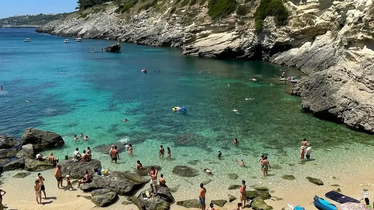 Spiaggia 100 gradini Mongerbino, con acque cristalline e scogliere rocciose. Persone nuotano e prendono il sole in una giornata estiva.