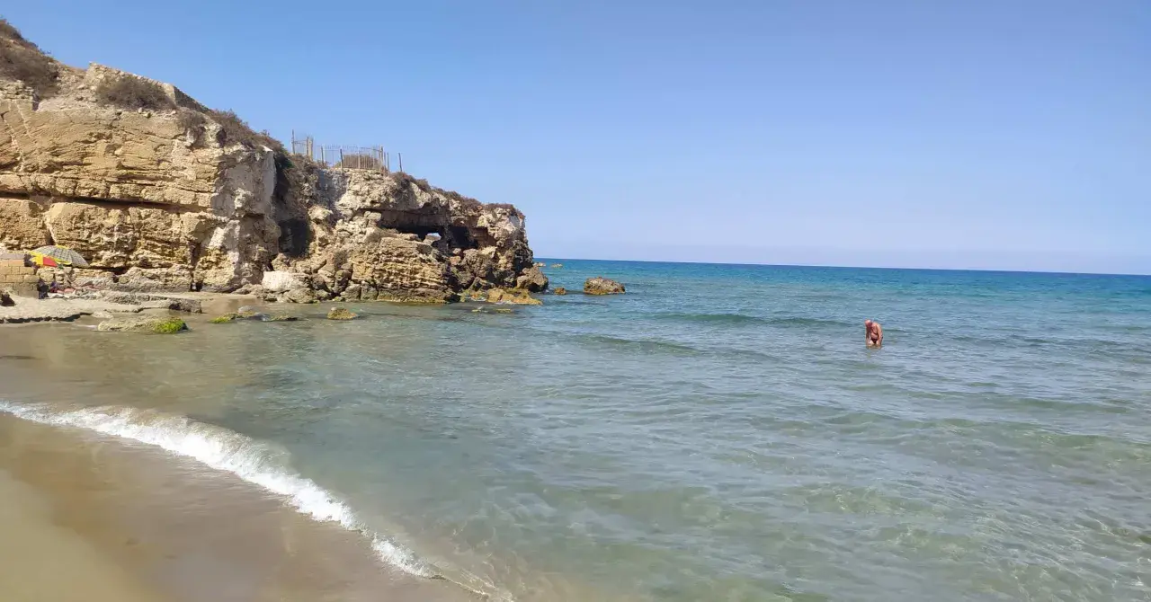 Spiaggia di Eloro con scogliere rocciose, mare azzurro e una persona che fa il bagno.