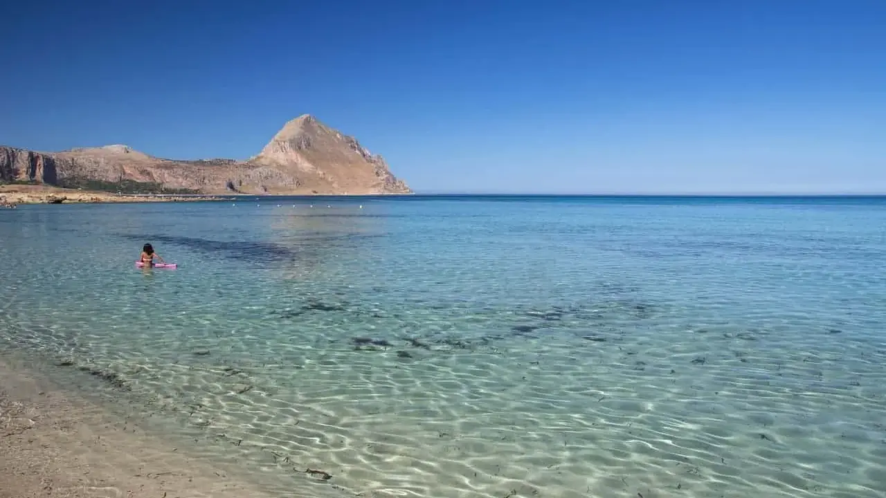 Bambina su un materassino rosa galleggia nelle acque cristalline della macari spiaggia, con montagne sullo sfondo.