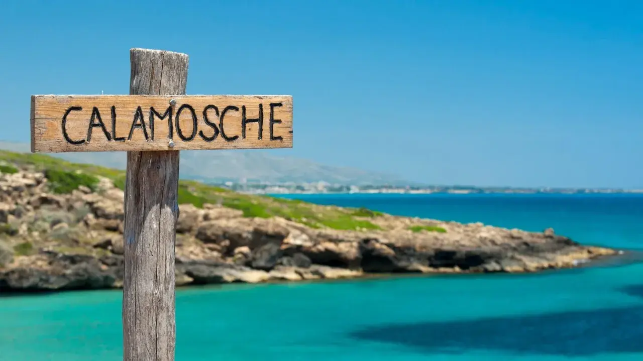 Cartello in legno "Calamosche Sicilia" su un palo, con mare turchese e costa rocciosa sullo sfondo.