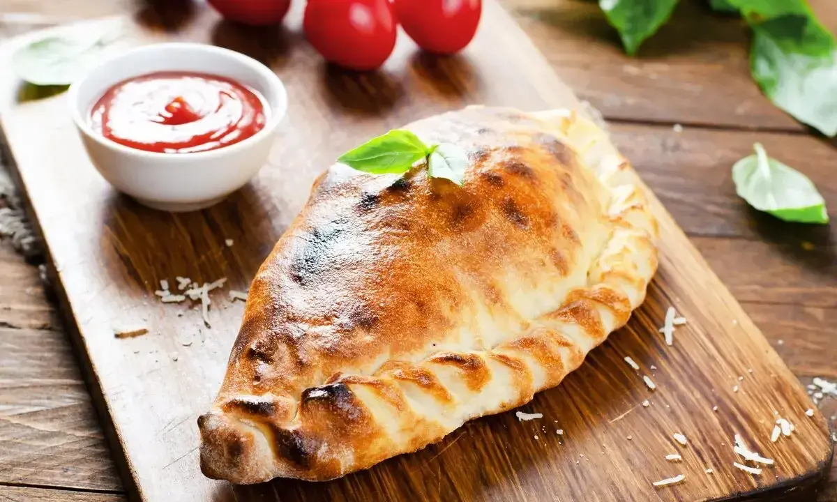 Un dorato cudduruni palermitano, guarnito con basilico, su tagliere di legno, con pomodorini e salsa di pomodoro.