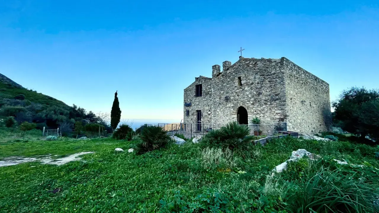 L'eremo di San Felice, un antico edificio in pietra con una croce sulla cima, si erge su una collina verdeggiante sotto un cielo azzurro.