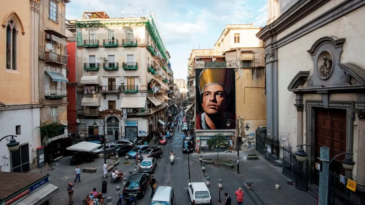 Un imponente murales di San Gennaro domina la scena in Via Forcella, Napoli. La strada è animata da auto, moto e persone.