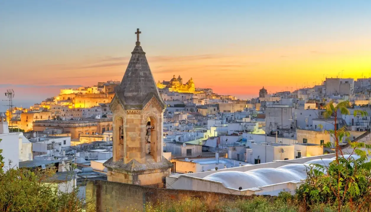 Campanile in primo piano, sullo sfondo la città bianca di Ostuni illuminata dal tramonto. Perfetto per scoprire cosa vedere a Ostuni in mezza giornata.