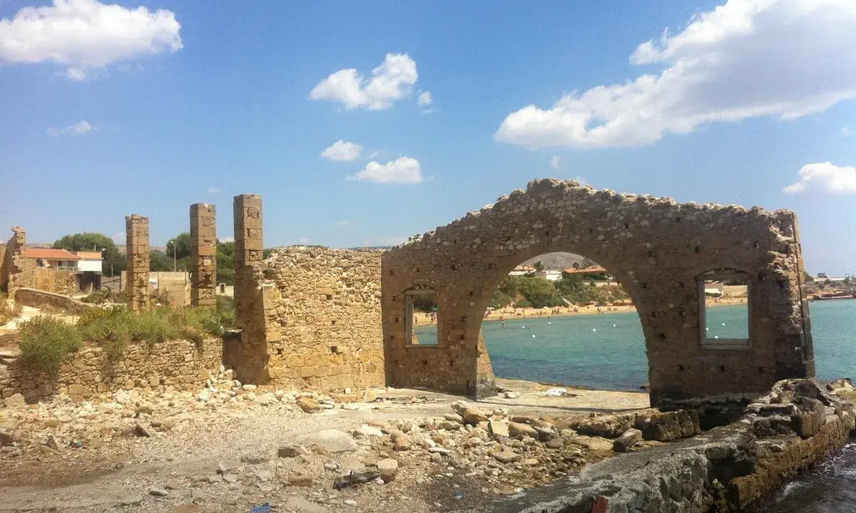 Rovine di una tonnara ad Avola, con un arco in pietra che si affaccia su una spiaggia affollata e il mare azzurro.