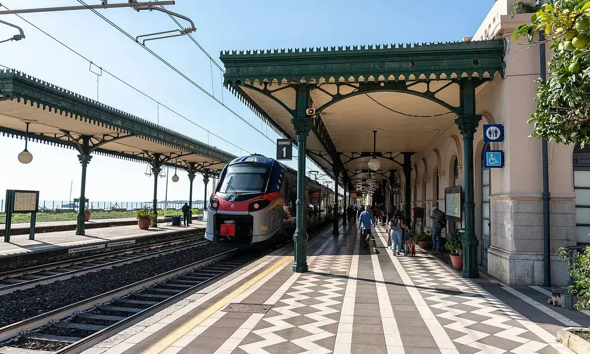 Treno in arrivo alla **stazione Taormina** sotto un portico verde. Persone attendono sul binario 1.