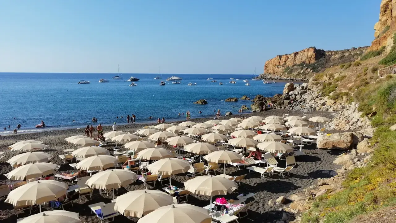 Spiaggia con ombrelloni e lettini, mare azzurro con barche e una scogliera. La torre conca domina il paesaggio.