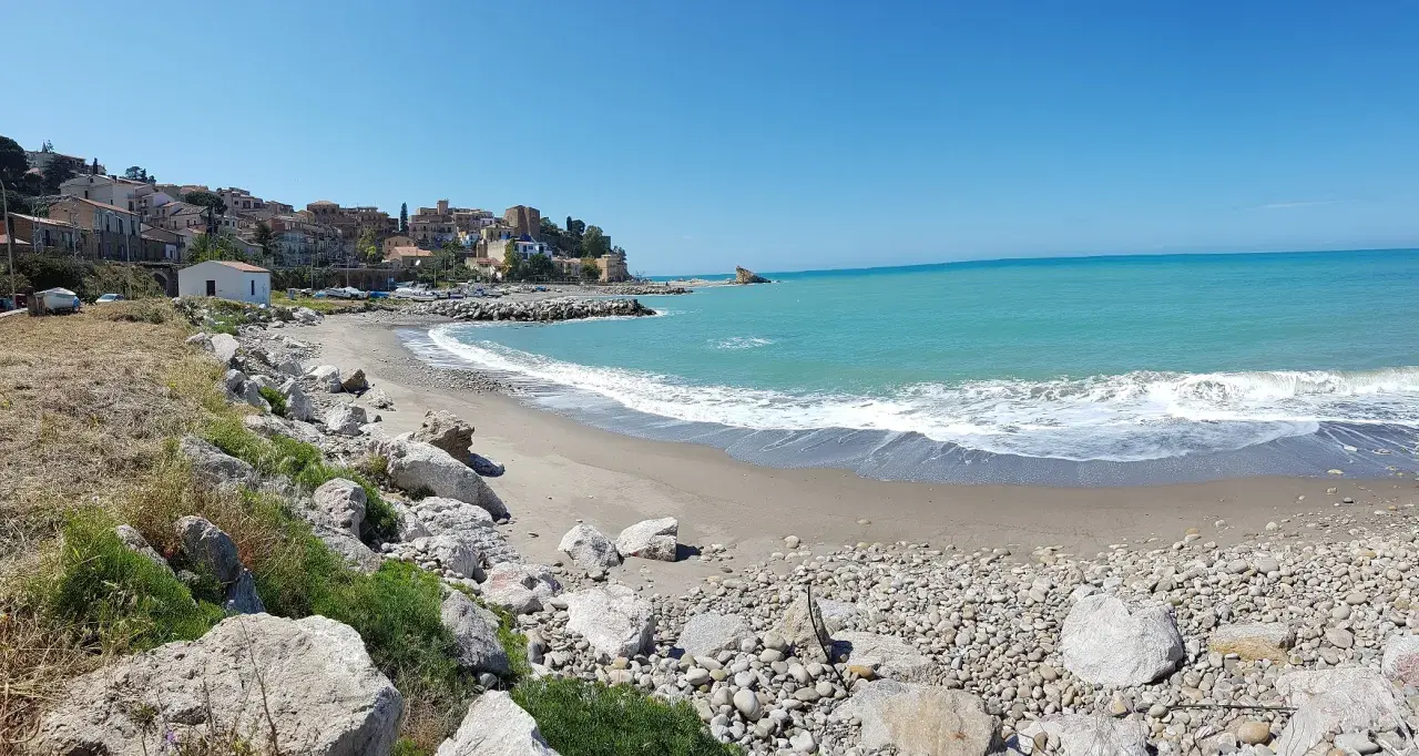 Spiaggia di Lampare a Tusa, con mare turchese, case colorate sul promontorio e rocce sulla riva.