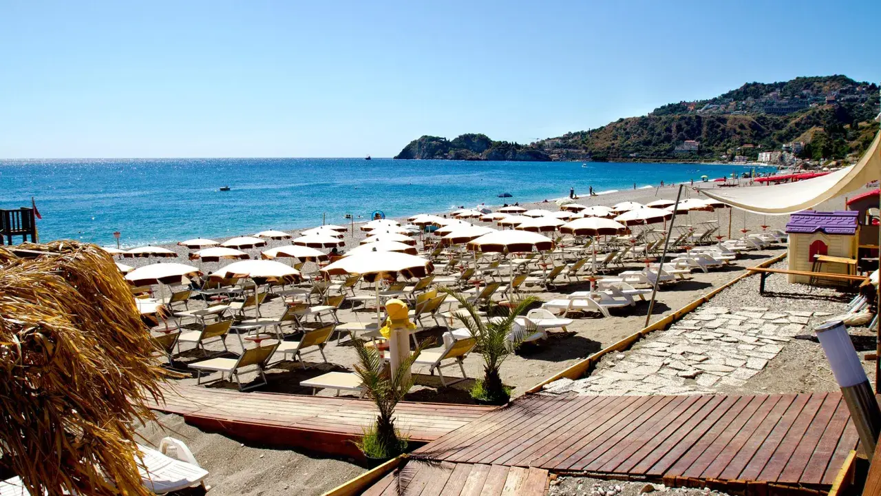 La spiaggia di Mazzeo, con file di ombrelloni bianchi e lettini, si estende verso il mare azzurro sotto un cielo limpido.