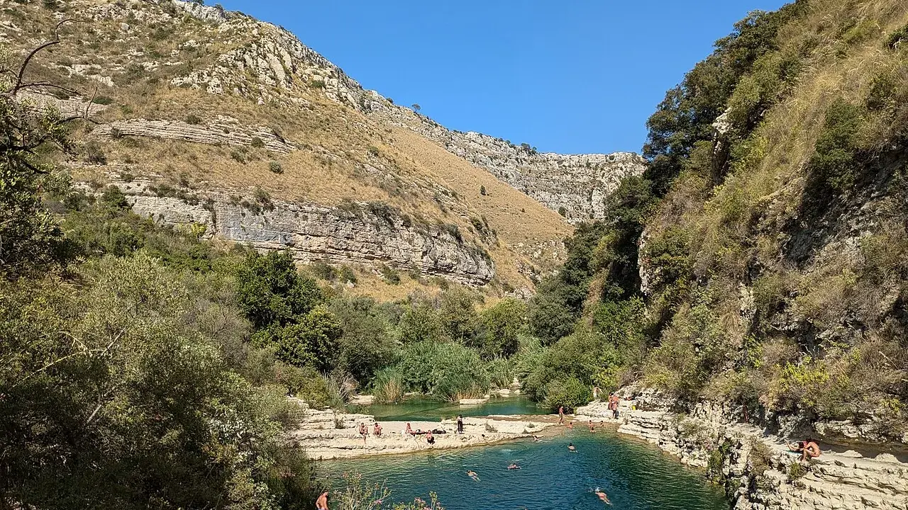 Gente si gode il sole e nuota nei laghetti d'Avola, circondati da rocce e vegetazione lussureggiante sotto un cielo azzurro.