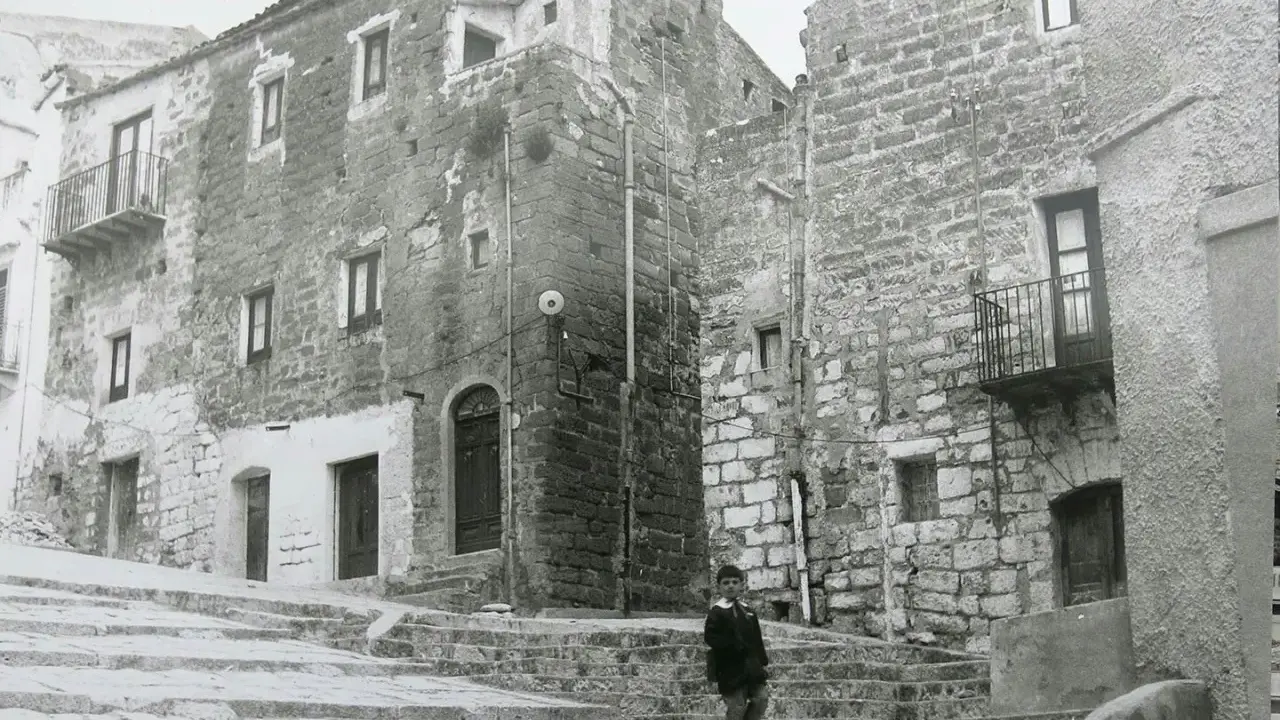 Un bambino cammina su una scalinata di pietra a Gibellina prima del terremoto. Edifici antichi in pietra la circondano.