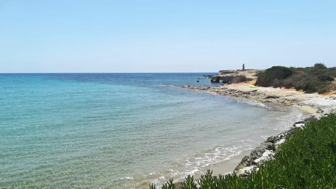 Acque cristalline e scogliere rocciose sulla spiaggia delle Formiche, Portopalo di Capo Passero. Un faro solitario veglia sul mare.