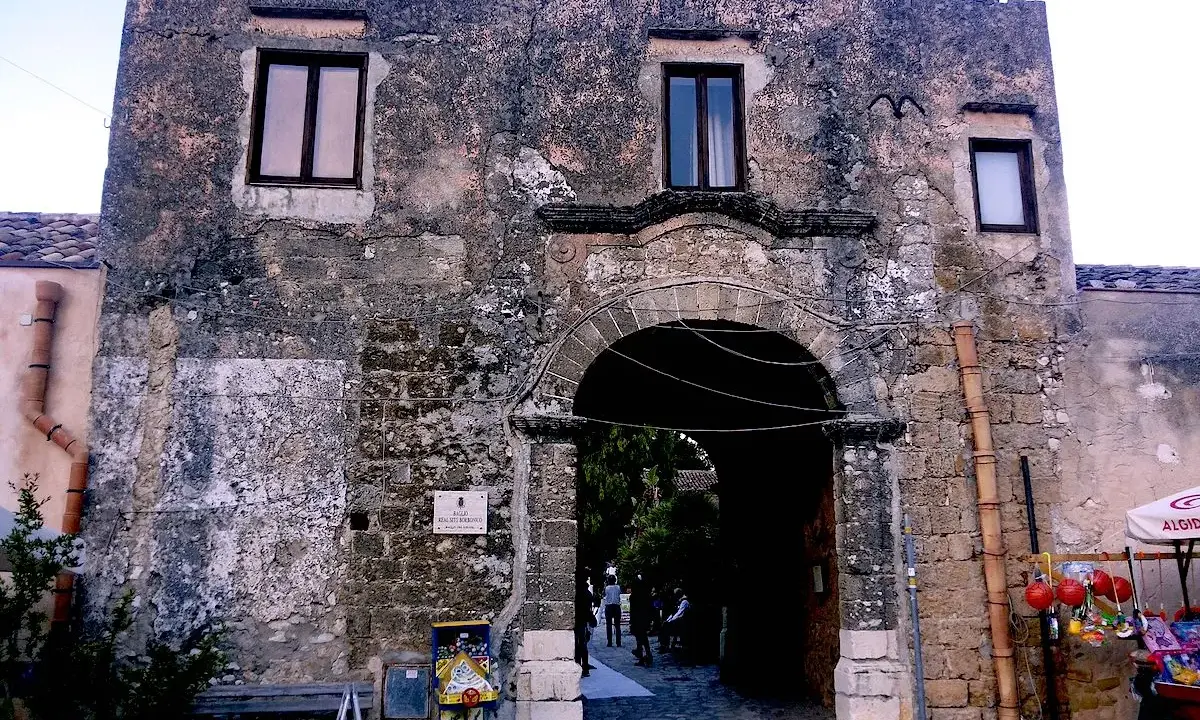 Ingresso di un baglio, con arco in pietra e muri antichi. Un piccolo chiosco vende souvenir.