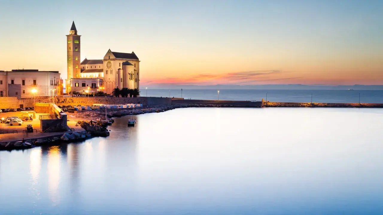 Tramonto sul porto di Trani, con la cattedrale illuminata che domina il lungomare. I vicoli del centro storico di Trani si intravedono tra gli edifici.