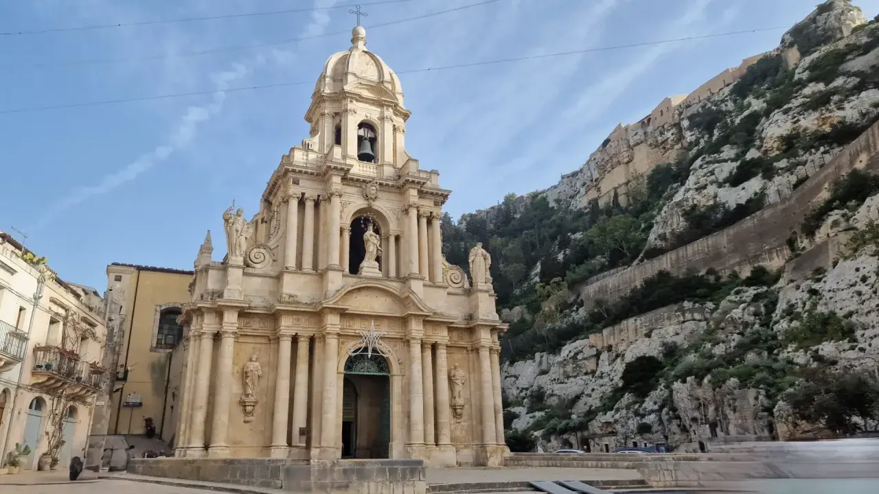 La chiesa barocca di San Giuseppe a Scicli, con il suo Cristo in gonnella, si erge maestosa contro una scogliera rocciosa.