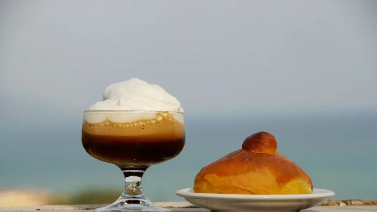 Un bicchiere di caffè freddo siciliano con panna e una brioche, con il mare sullo sfondo.