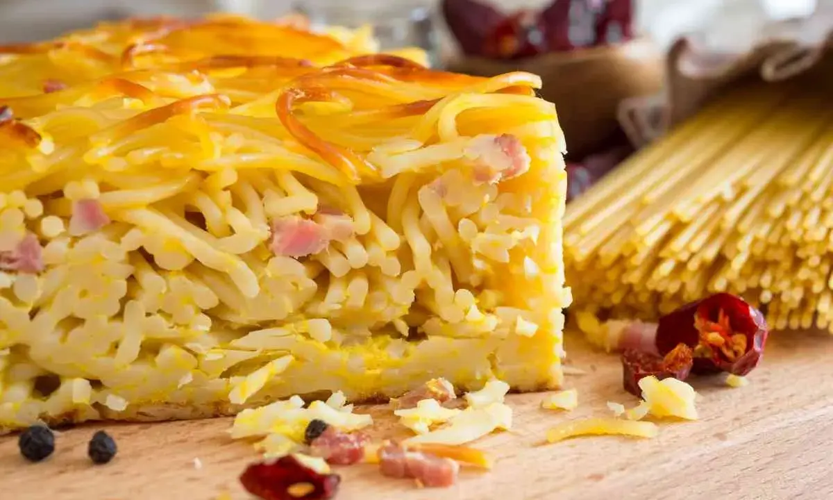 Fetta di spaghetti al forno, uno dei piatti tipici napoletani, con pancetta e formaggio. Sullo sfondo, spaghetti crudi e peperoncini.