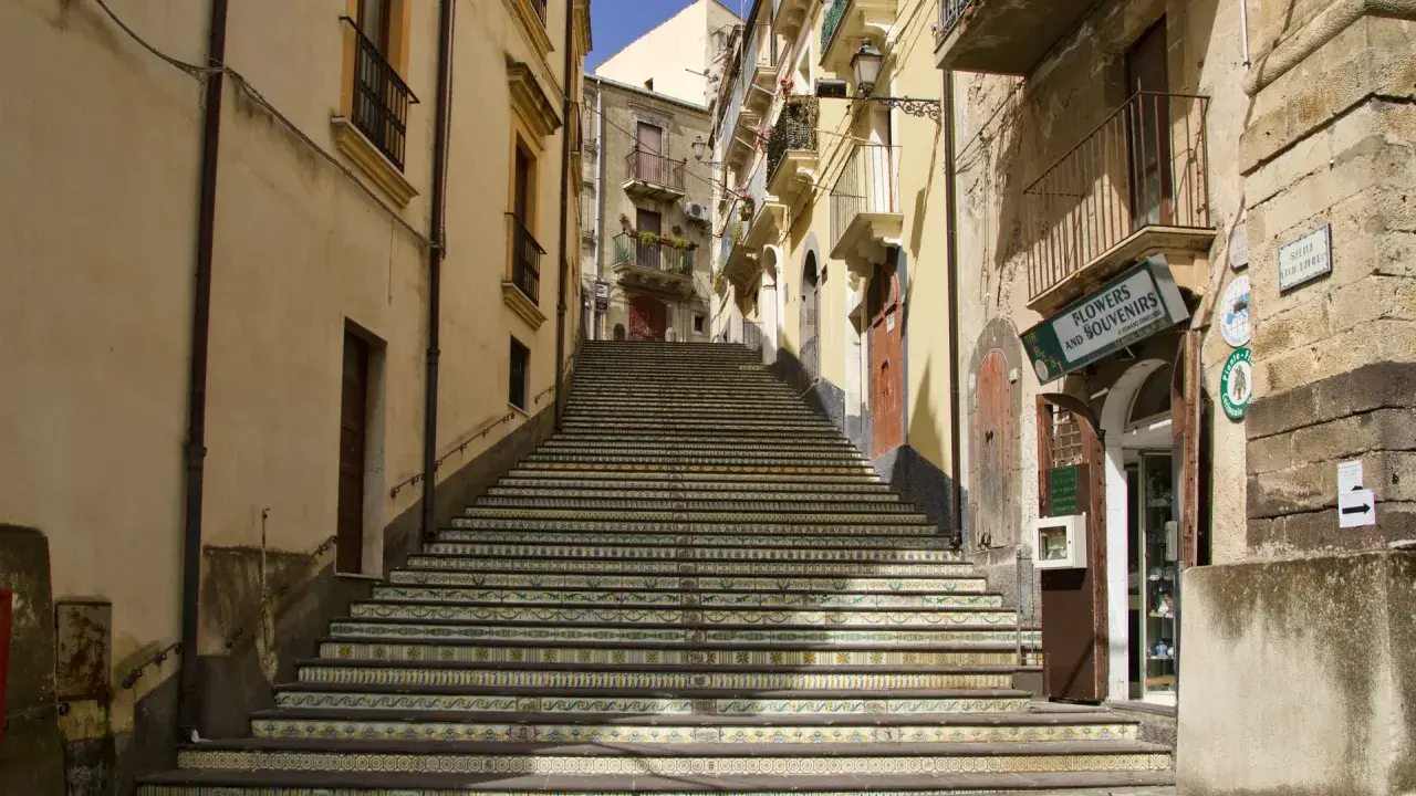 Una scalinata decorata con maioliche colorate sale tra edifici storici. Un negozio di fiori e souvenir invita a scoprire cosa vedere a Vizzini.