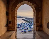 Arco antico incornicia la spiaggia di Cefalù con ombrelloni blu e lettini, vista sul mare e sul molo affollato.