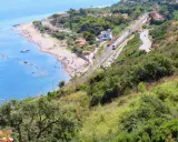 Spiaggia turchina finale di Pollina, con bagnanti e barche. Sopra, una ferrovia e una strada serpeggiano tra la vegetazione lussureggiante.