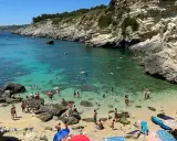 Spiaggia 100 gradini Mongerbino, con acque cristalline e scogliere rocciose. Persone nuotano e prendono il sole in una giornata estiva.