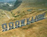 Aerial view of the "HOLLYWOOD" sign in Palermo, Italy, set against a dry, hilly landscape.
