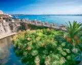 Siracusa, con la sua etimologia legata all'acqua, mostra papiri rigogliosi che galleggiano in un canale, con il mare azzurro e barche sullo sfondo.