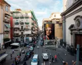 Un imponente murales di San Gennaro domina la scena in Via Forcella, Napoli. La strada è animata da auto, moto e persone.