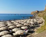 Spiaggia con ombrelloni e lettini, mare azzurro con barche e una scogliera. La torre conca domina il paesaggio.
