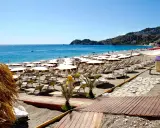 La spiaggia di Mazzeo, con file di ombrelloni bianchi e lettini, si estende verso il mare azzurro sotto un cielo limpido.