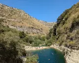 Gente si gode il sole e nuota nei laghetti d'Avola, circondati da rocce e vegetazione lussureggiante sotto un cielo azzurro.