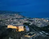 La rocca licata domina la città illuminata al crepuscolo, con il mare blu sullo sfondo.