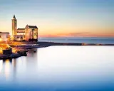 Tramonto sul porto di Trani, con la cattedrale illuminata che domina il lungomare. I vicoli del centro storico di Trani si intravedono tra gli edifici.