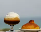 Un bicchiere di caffè freddo siciliano con panna e una brioche, con il mare sullo sfondo.