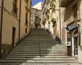 Una scalinata decorata con maioliche colorate sale tra edifici storici. Un negozio di fiori e souvenir invita a scoprire cosa vedere a Vizzini.