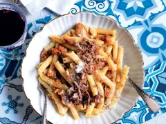 Piatto di pasta con ragù di carne, un classico dei piatti napoletani antichi, servito su un piatto bianco con bordo dorato, accanto a un bicchiere di vino rosso.