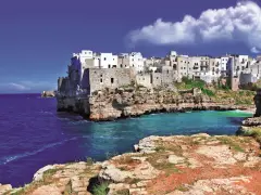 Panoramica di Polignano a Mare, con le case bianche aggrappate alle scogliere a picco sul mare blu intenso. La **distanza Locorotondo Polignano a Mare** è breve per godersi questo spettacolo.