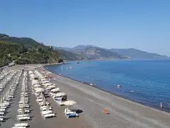 Spiaggia Torre Conca: file di lettini e ombrelloni su ghiaia scura, mare azzurro e colline verdi sullo sfondo.