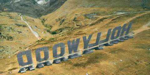 Aerial view of the "HOLLYWOOD" sign in Palermo, Italy, set against a dry, hilly landscape.