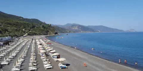 Spiaggia Torre Conca: file di lettini e ombrelloni su ghiaia scura, mare azzurro e colline verdi sullo sfondo.