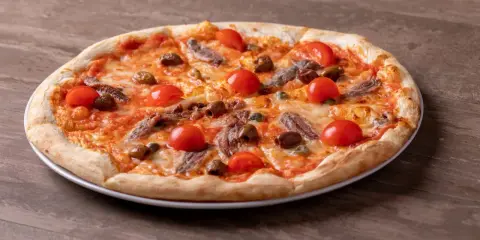 Pizza fuata siciliana con pomodorini, acciughe, olive e capperi su un piatto bianco, su un tavolo di legno.