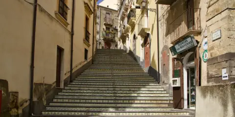 Una scalinata decorata con maioliche colorate sale tra edifici storici. Un negozio di fiori e souvenir invita a scoprire cosa vedere a Vizzini.