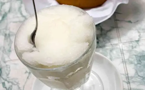 Un bicchiere colmo di granita al gelsomino, con un cucchiaino che affonda nella sua consistenza ghiacciata. Perfetta per una pausa rinfrescante.