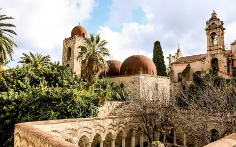 Le iconiche cupole rosse di Palermo si stagliano tra palme e vegetazione lussureggiante, un'antica bellezza sotto un cielo nuvoloso.