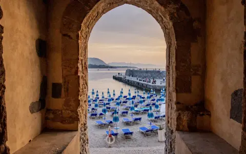 Arco antico incornicia la spiaggia di Cefalù con ombrelloni blu e lettini, vista sul mare e sul molo affollato.
